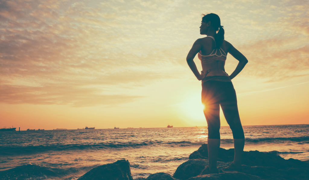Gesundheit und Vorsorge: Frau steht mit dem Rücken zur Kamera im Sportoutfit im Sonnenuntergang auf Steinen an der Küste und blickt aufs Meer