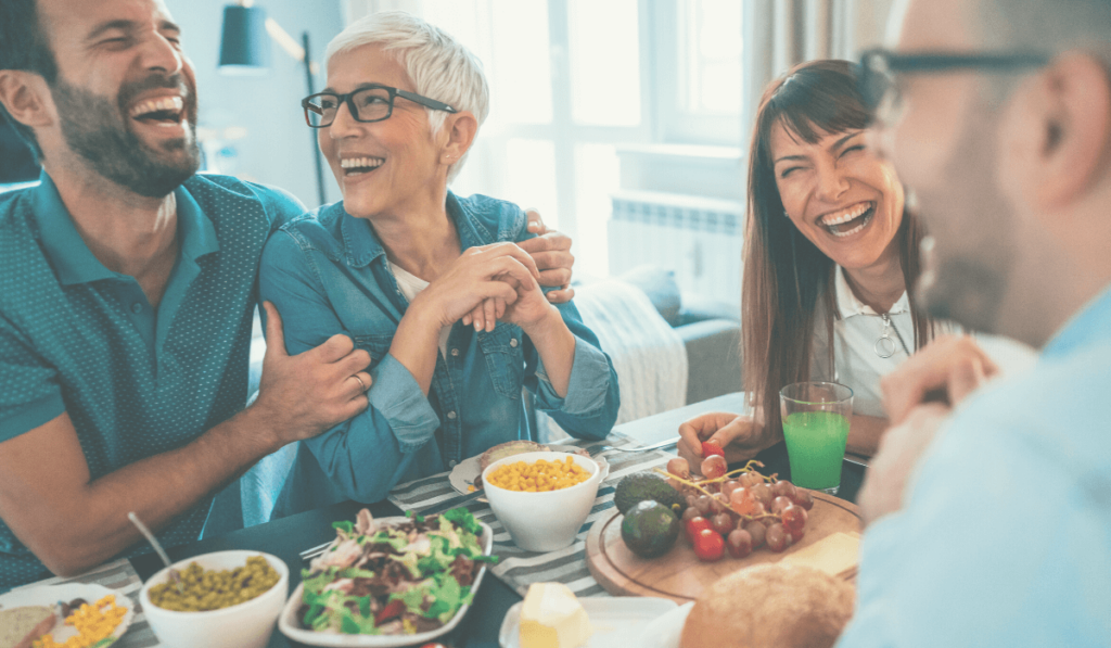 Zwei Männer und zwei Frauen unterschiedlichen Alters sitzen am Esstisch mit gesundem Essen und lachen herzlich